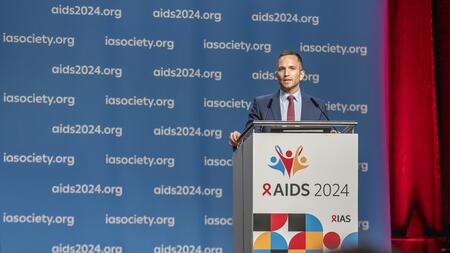 Prof. Christoph Spinner steht in einem dunkelblauen Anzug mit roter Krawatte an einem Rednerpult und spricht in ein Mikrofon. Das Rednerpult trägt das Logo und die Aufschrift "AIDS 2024". Im Hintergrund ist eine blaue Wand zu sehen, auf der wiederholt die Texte "aids2024.org" und "iasociety.org" gedruckt sind. Rechts im Bild ist ein roter Vorhang sichtbar.
