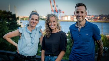 The picture shows the Infektiopod team of three standing in front of a harbour landscape in Hamburg. All three people are smiling at the camera. On the left is Annette Hennigs with a blue sleeveless top, in the centre is Elena Terhalle with curly, shoulder-length hair and a black T-shirt, and on the right is Till Koch with short hair, a blue polo shirt and tattooed arms. In the background you can see cranes and buildings on the waterfront.